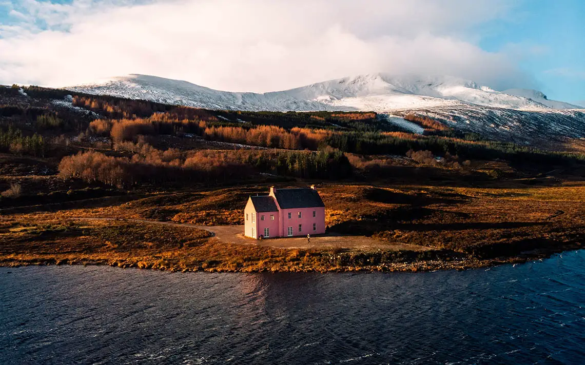 The Pink House in the Scottish Highlands with a backdrop of snow-capped mountains.